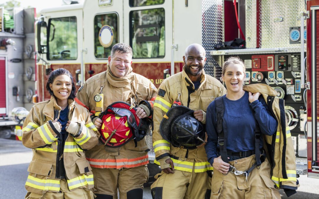 A multi-ethnic group of four firefighters standing together in front of fire engines, wearing fire protection suits and holding helmets. Two of them are women. They are looking at the camera, smiling.